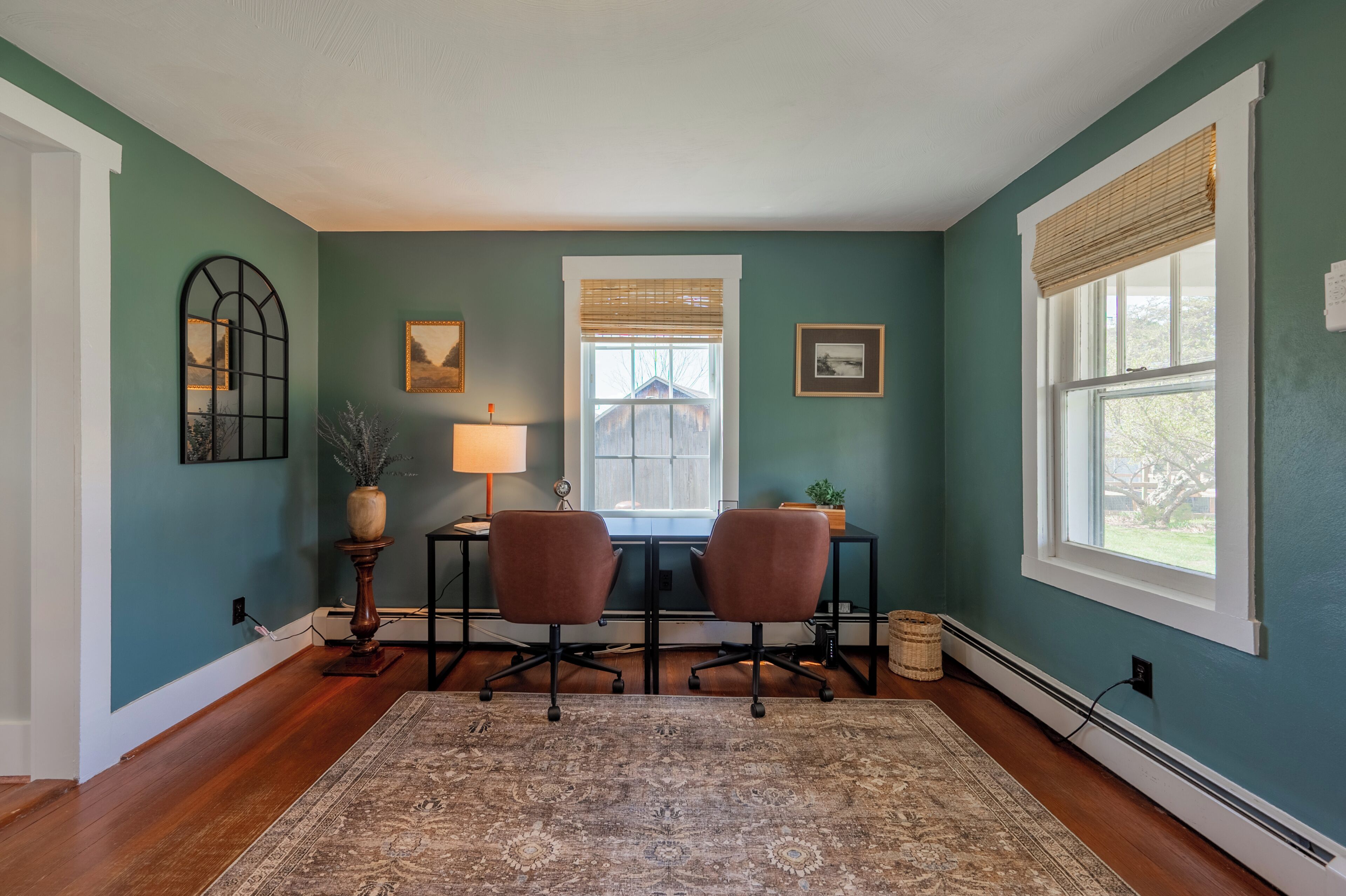 Een kamer met blauwgroene wanden, een houten vloer, een bureau met twee stoelen, een lamp en lijstjes met afbeeldingen.