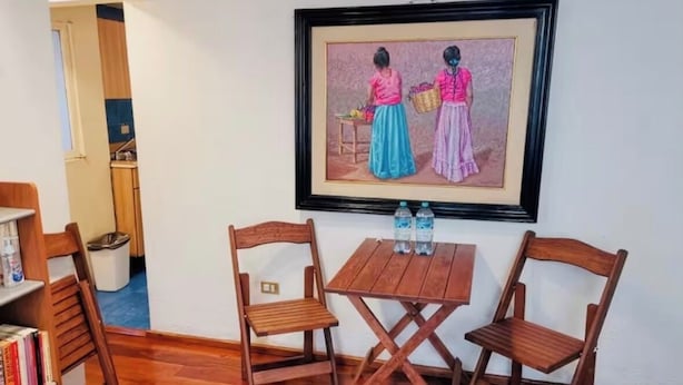Una stanza con pavimento in legno, un quadro incorniciato, due sedie di legno, un tavolino e una libreria.
