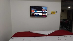 A bed with a red headboard and a white floral bedspread, a flat-screen TV mounted on the wall displaying a streaming service interface, and a yellow power strip on the wall.