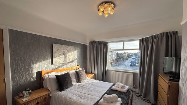 Habitación de hotel con cama, mesita de noche de madera, televisor, ventana con cortinas y radiador.