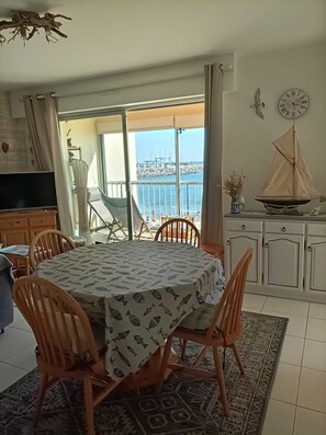 A dining area with a table set for four, chairs, a television, and a view of the sea through large windows. - APARTMENT FROND DE MER (La Turballe)