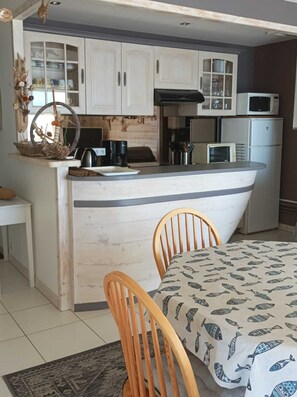 A compact kitchen with white cabinets, a microwave, a refrigerator, and a small dining area with a table and chairs. - APARTMENT FROND DE MER (La Turballe)