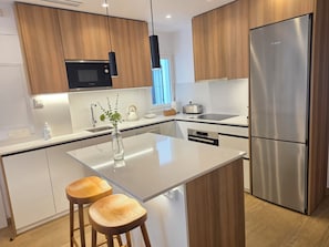 Une cuisine moderne avec des meubles en bois, un réfrigérateur en acier inoxydable, un four à micro-ondes Bosch, un plan de travail blanc et deux tabourets en bois.