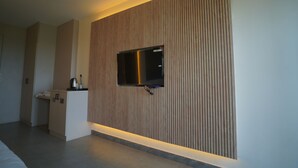 A modern hotel room with a flat-screen TV mounted on a wooden panel wall, a small kitchenette, and a bed.