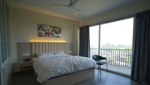 A bedroom with a bed, a nightstand, a chair, a ceiling fan, a painting, and a large window with curtains.