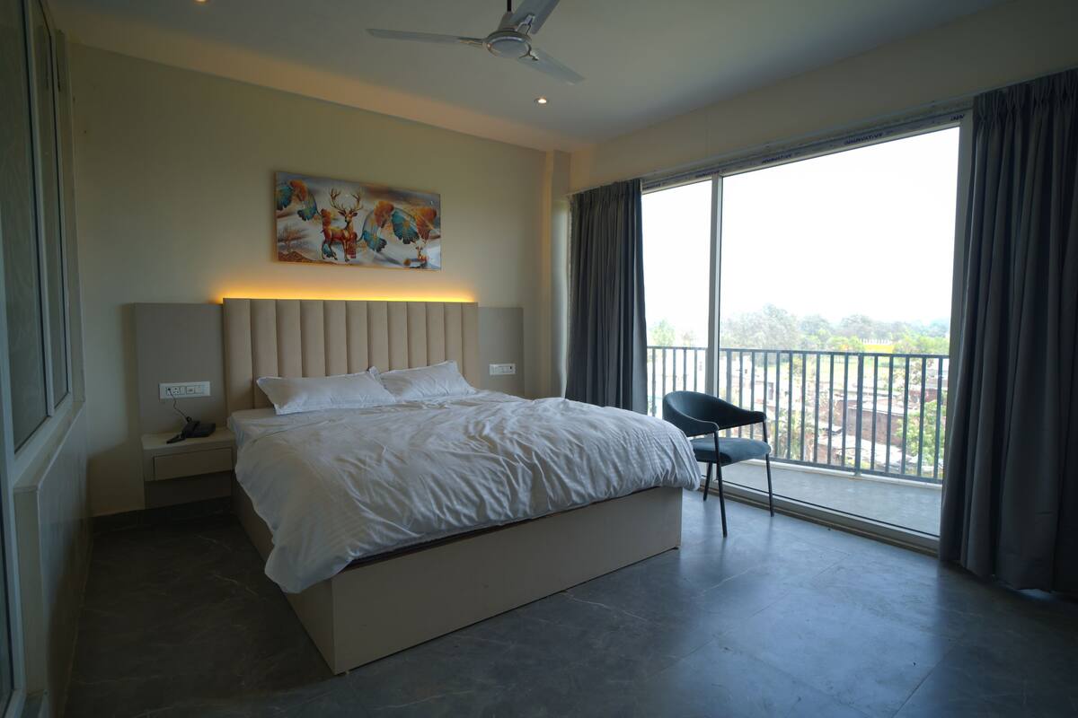 Une chambre à coucher comprenant un lit, une table de chevet, une chaise, un ventilateur de plafond, un tableau et une grande fenêtre avec des rideaux.