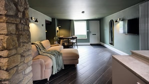 Un salon avec un mur en pierre, un canapé beige, une table à manger avec des chaises et un téléviseur à écran plat fixé au mur.