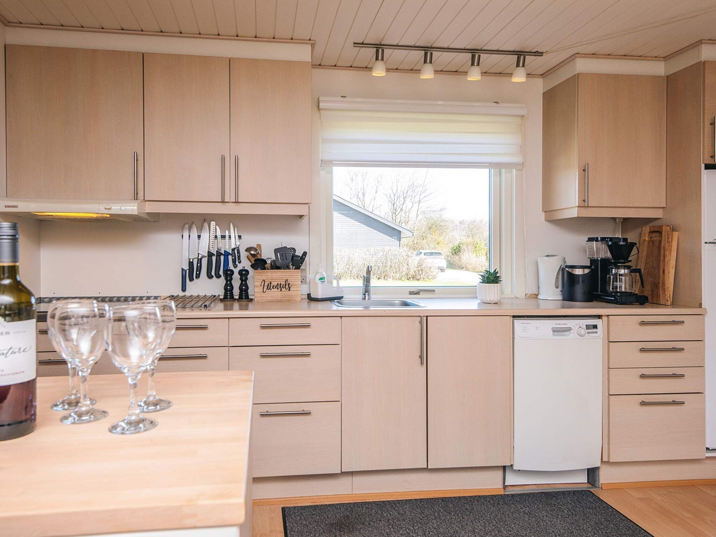 A modern kitchen with light wood cabinets, a built-in dishwasher, and a window with a view of the outside.