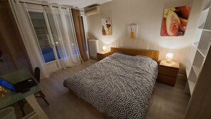 Ein Hotelzimmer mit Bett, Schreibtisch, Stuhl, Fenster mit Vorhängen, Heizkörper und Wandbildern.