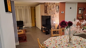 Ein Wohnzimmer mit Fernseher, einer Couch, einem Esstisch mit Obstmuster-Tischdecke und einer Wand mit Holzstruktur.
