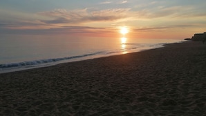 Uma praia com o sol se pondo no horizonte, mar calmo e falésias ao longe.