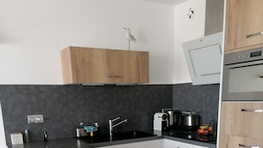Uma cozinha moderna com forno embutido, pia e bancada com diversos eletrodomésticos. Há dois bancos e uma pequena mesa.