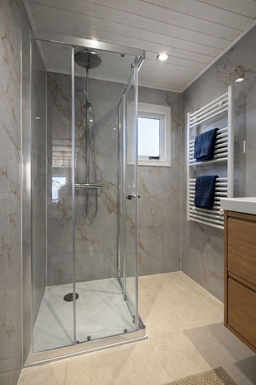 Une douche fermée par des parois en verre, dotée d’un pommeau de douche à main, d’une petite fenêtre et d’un porte-serviettes avec des serviettes bleues.