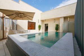 A swimming pool with a stone edge, a sun umbrella, and potted plants.
