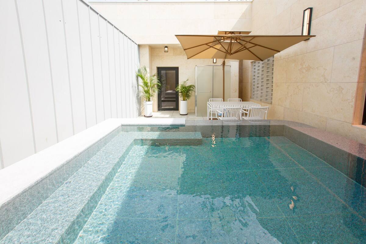 Ein Swimmingpool mit weißen Stühlen und einem Tisch, umgeben von Mauern und Topfpflanzen.