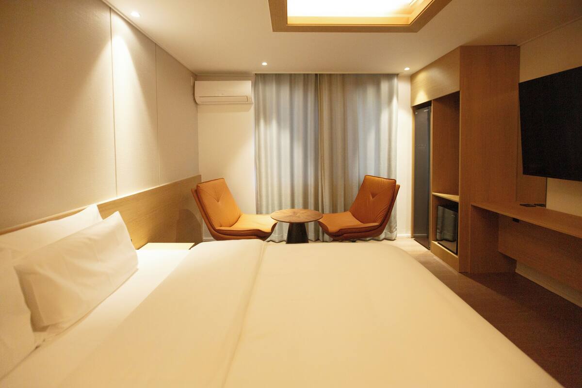 Ein Hotelzimmer mit einem Bett, zwei Sesseln, einem kleinen Tisch, einem Fernseher und einem Fenster mit Vorhängen.