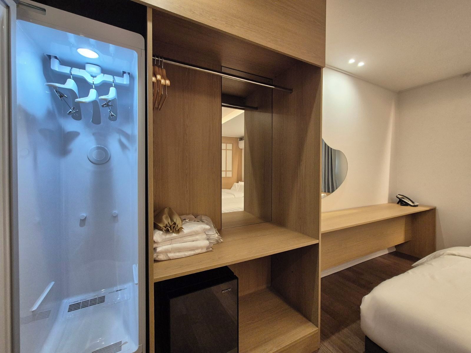 A hotel room with a built-in shower, a mirror, a bedside table, and a neatly folded towel on the floor.
