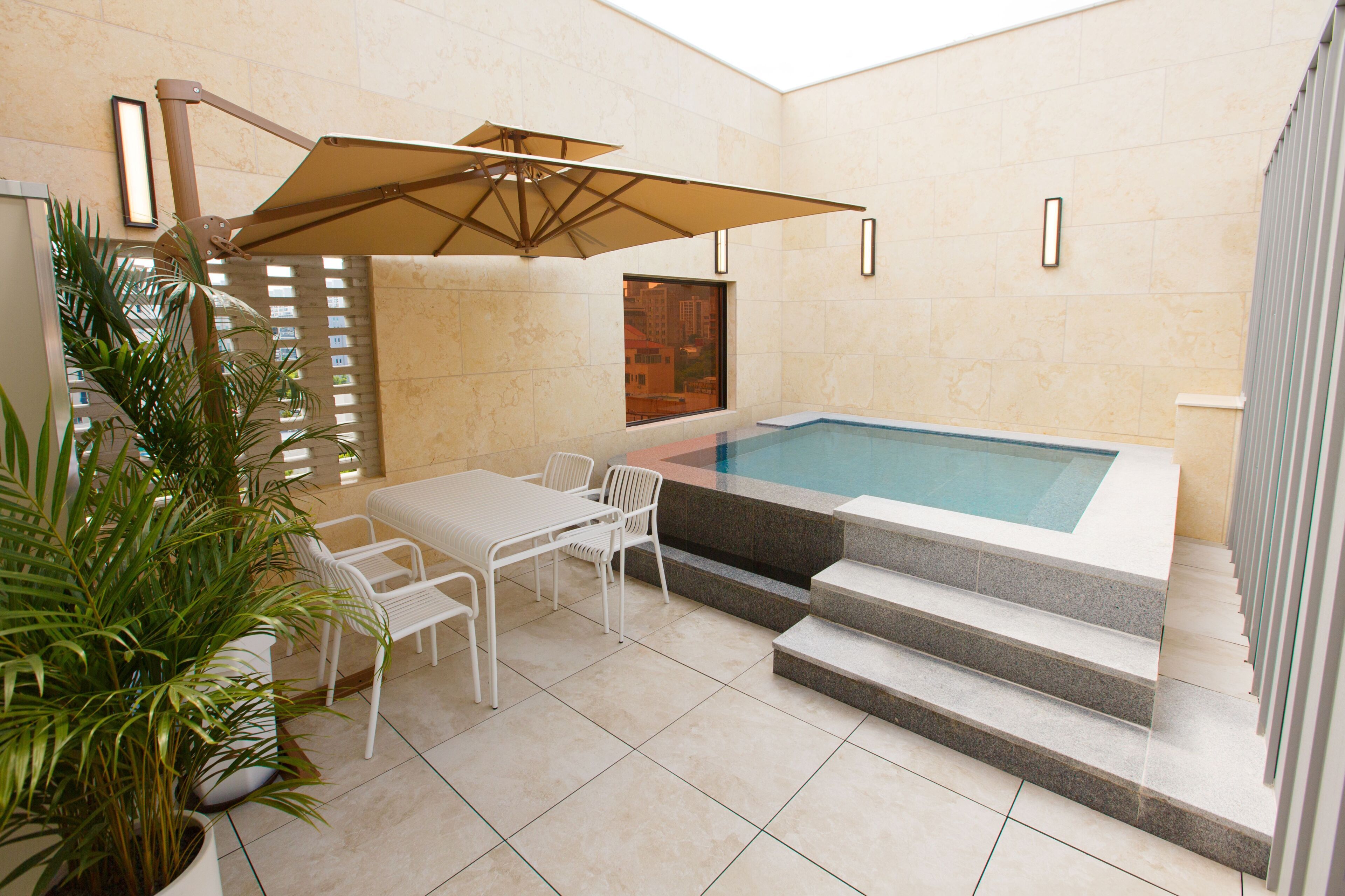 A modern indoor pool area with a hot tub, white chairs, a table, and a large umbrella.