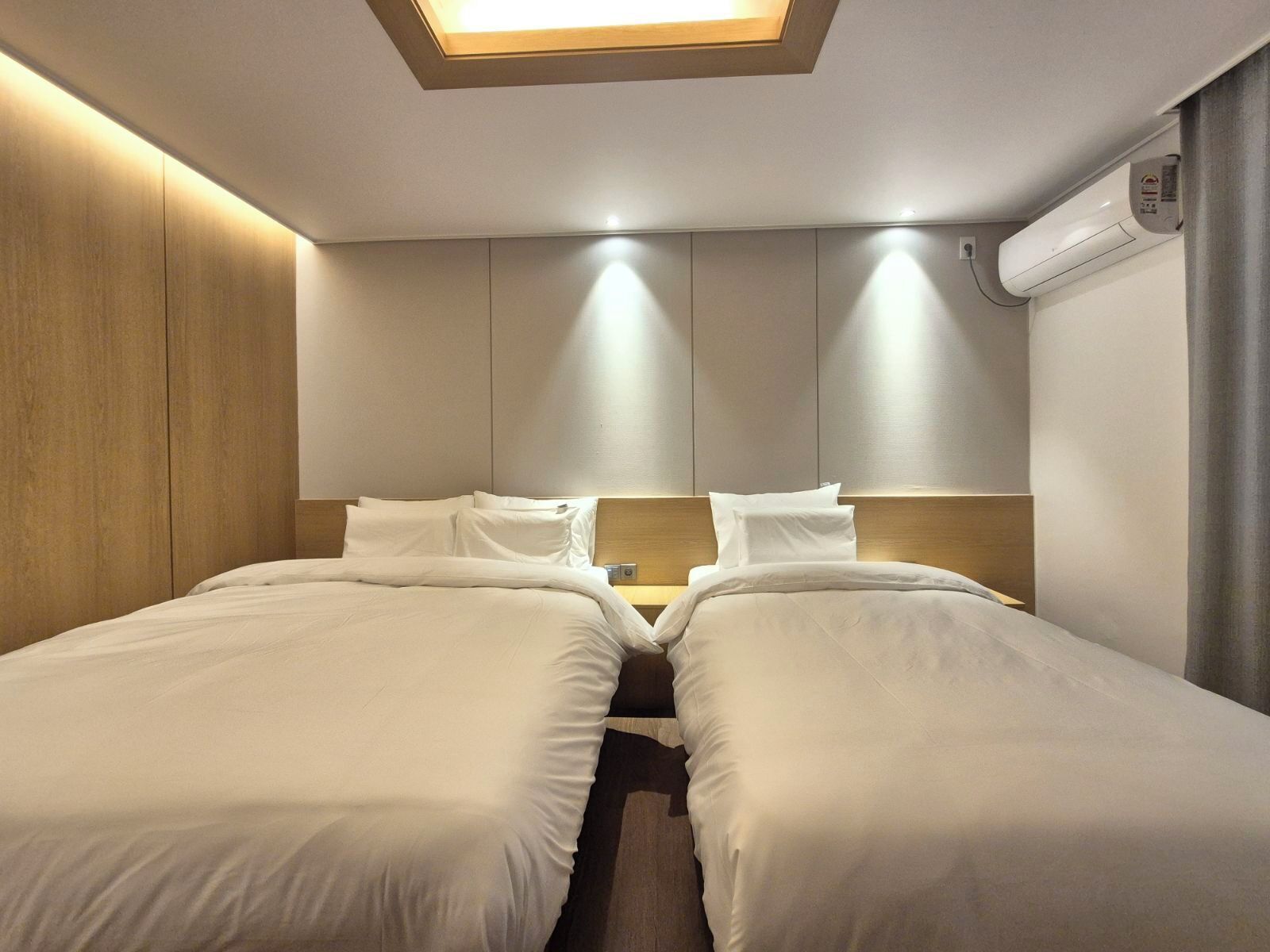 A hotel room with two beds, a skylight, a wall-mounted air conditioner, and a wooden accent wall.