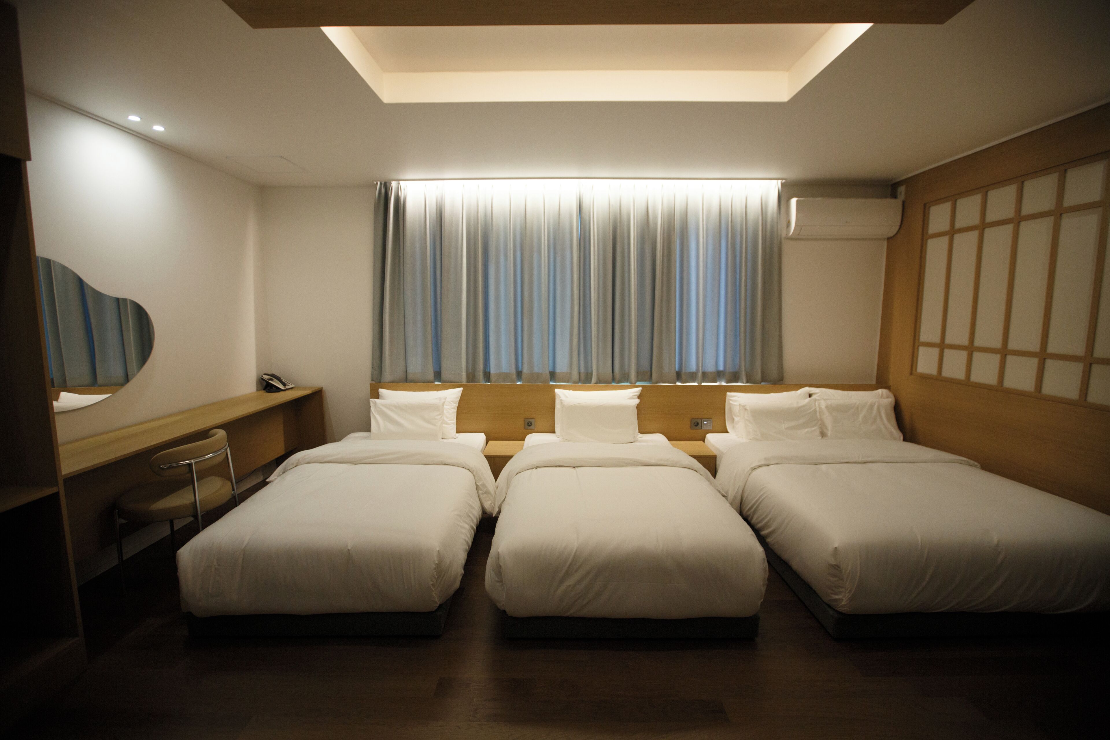 Habitación de hotel con tres camas, un escritorio, una silla, un ventanal con cortinas y un tragaluz.