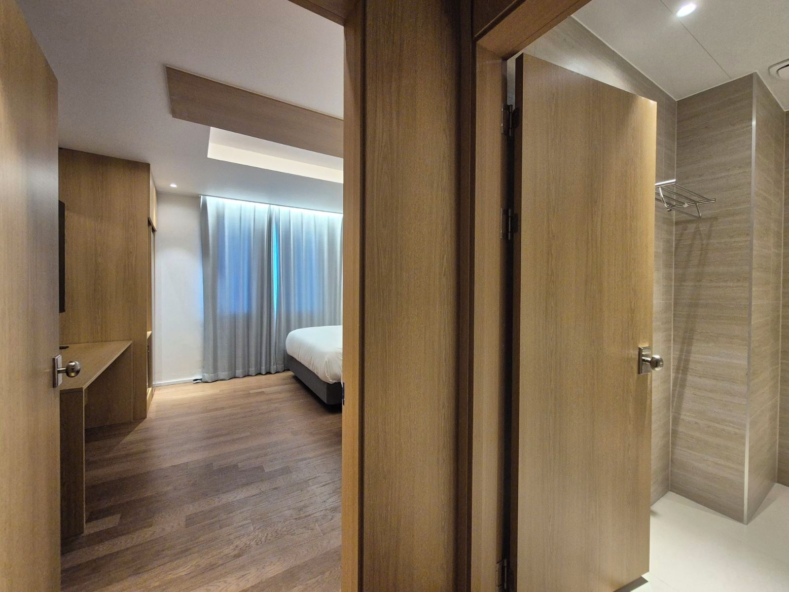 A hotel room with a wooden floor, a bed, a shower area, and a window with curtains.