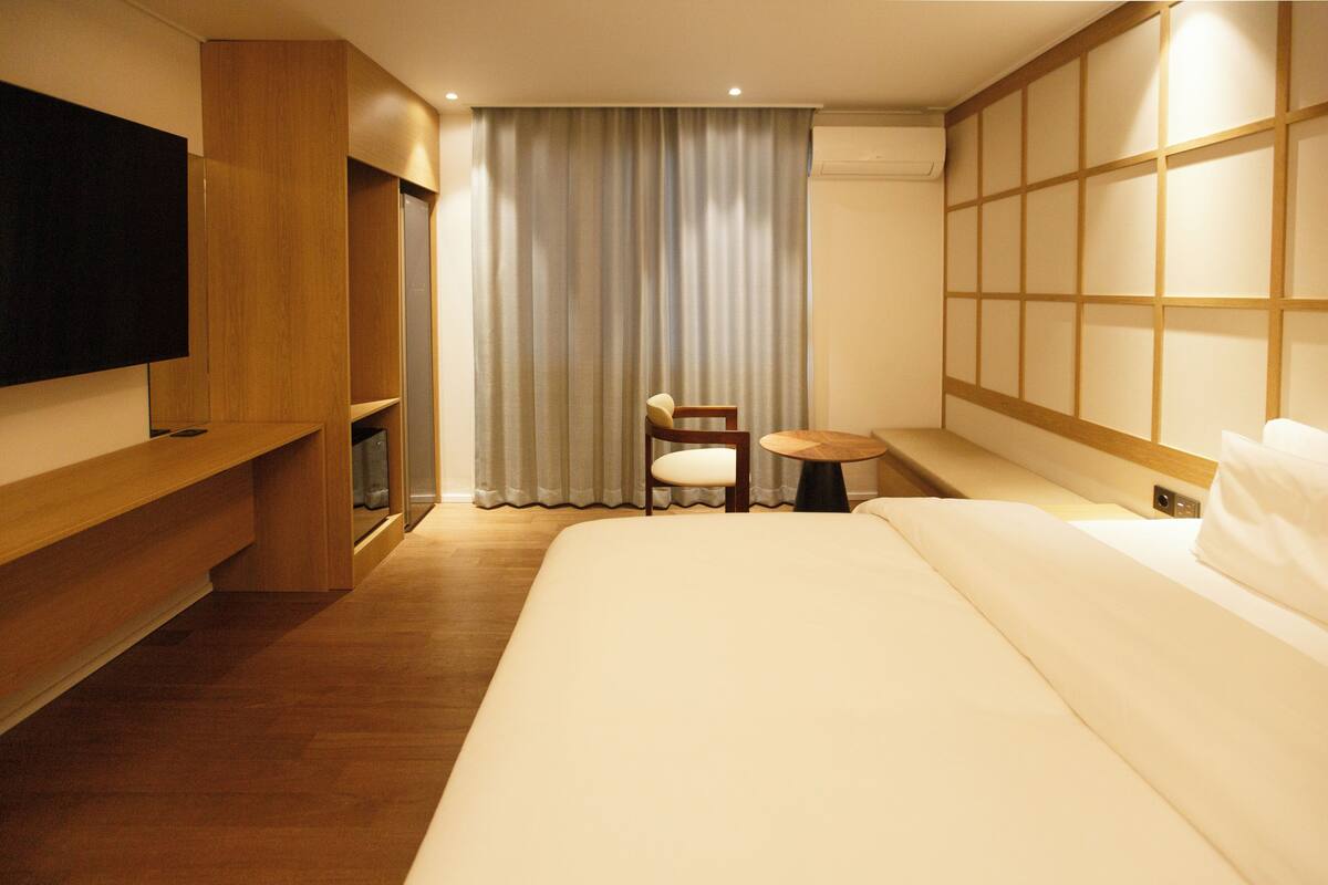 Ein Hotelzimmer mit einem großen Bett, einem an der Wand befestigten Fernseher, einem Holztisch, einem Stuhl und einem kleinen Tisch.