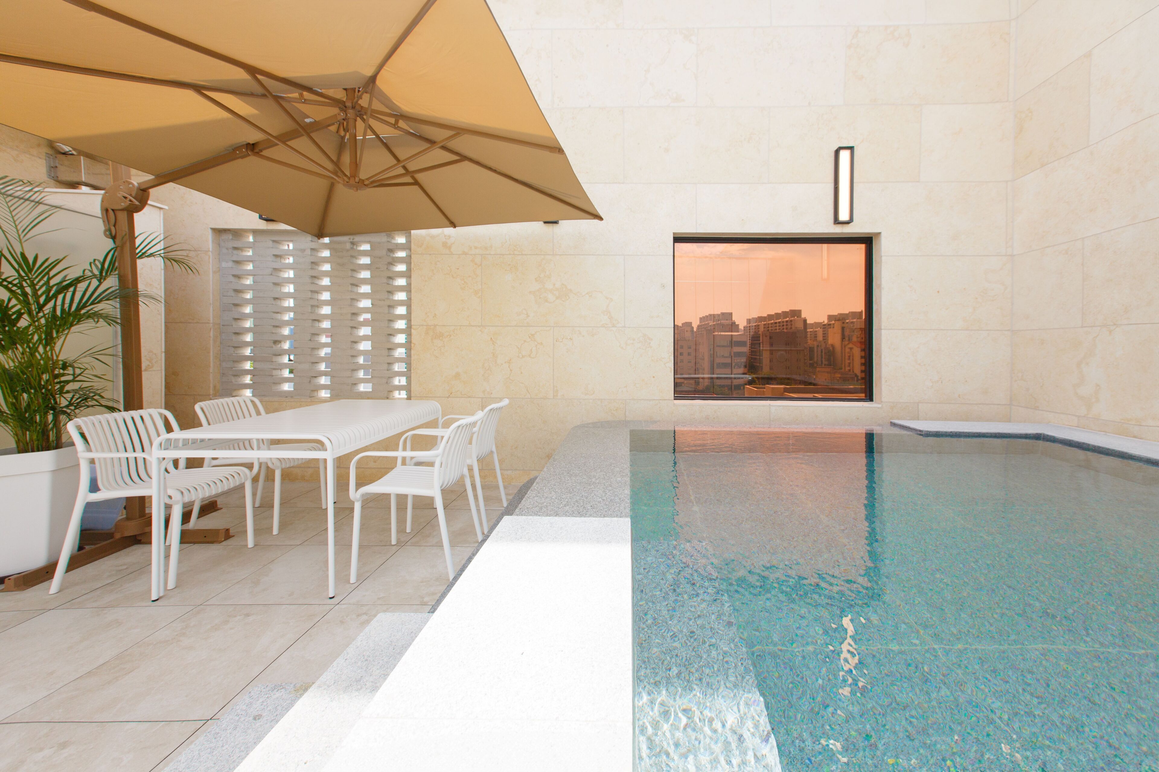 Una piscina interior con vistas a un paisaje urbano a través de una ventana, muebles blancos para exteriores y un paraguas grande.