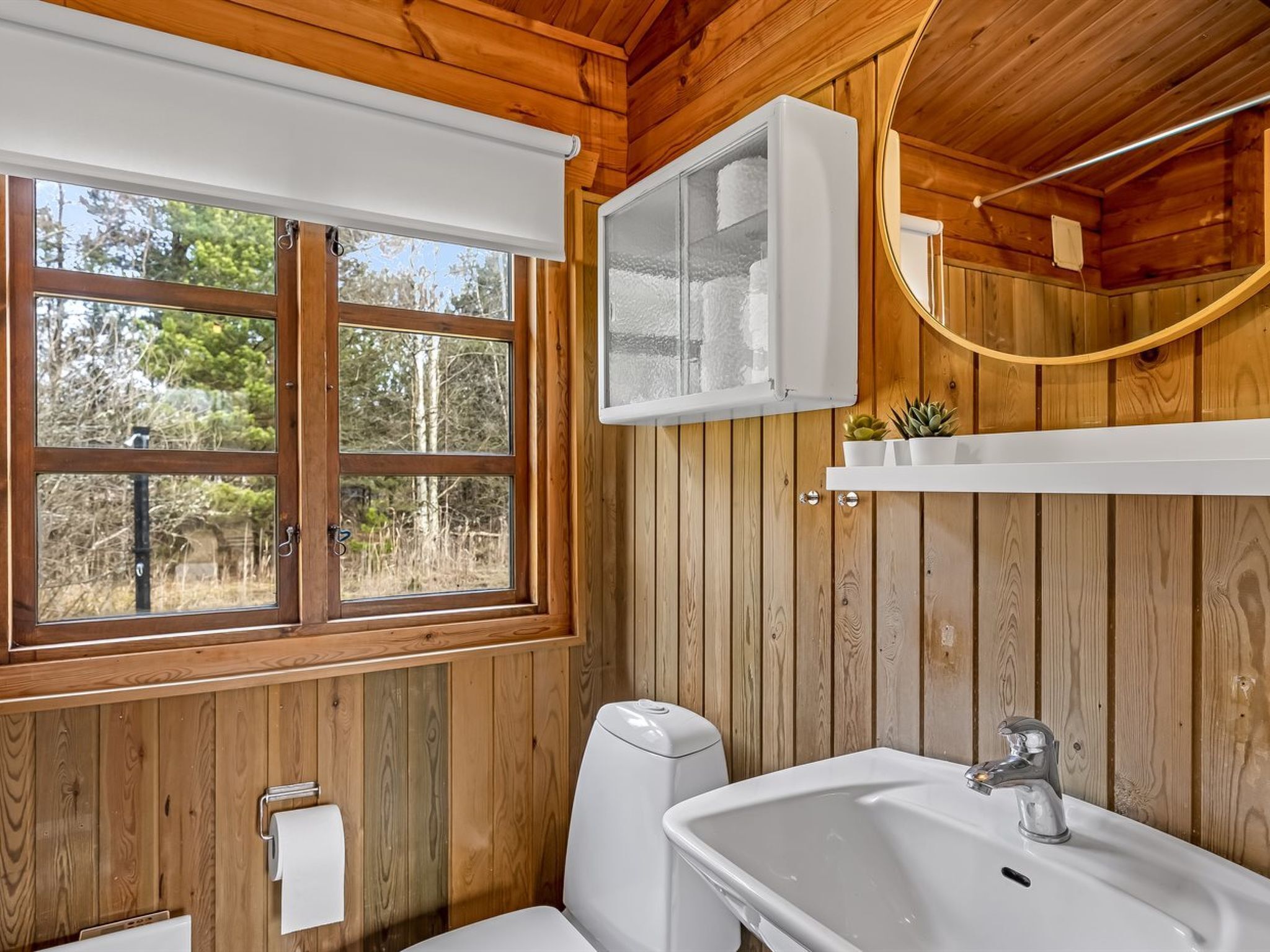 A wooden cabin bathroom with a toilet, sink, and window.