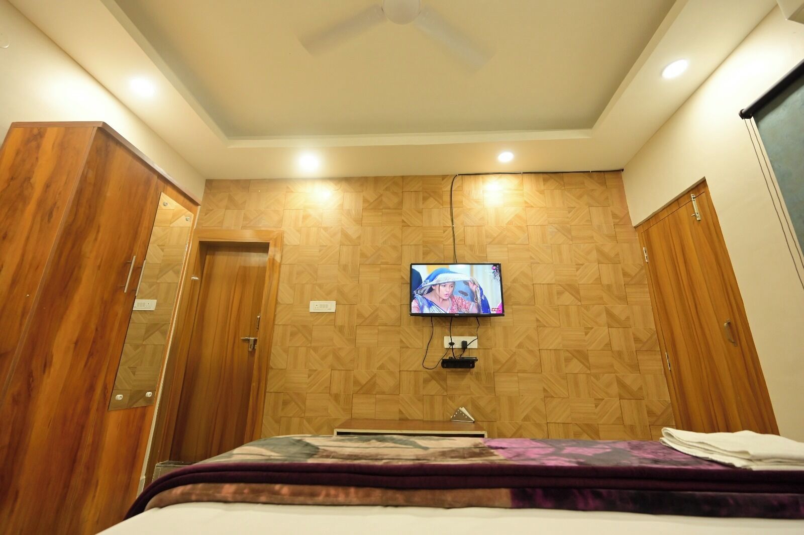 A hotel room with a bed, a television mounted on the wall, wooden paneling, and a ceiling fan.