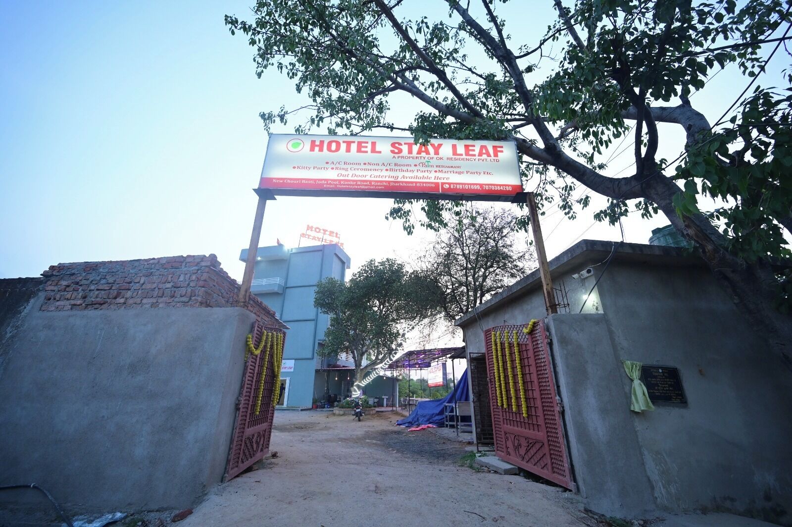 Entrada de hotel con un cartel que indica el nombre 'HOTEL STAY LEAF' y servicios como habitaciones con aire acondicionado, habitaciones sin aire acondicionado y diversas opciones para eventos.