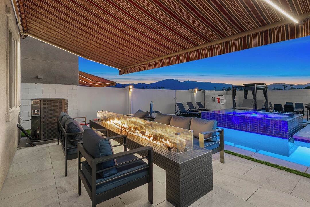 An outdoor patio with a fire feature, seating, a pool with blue lighting, and a view of mountains.