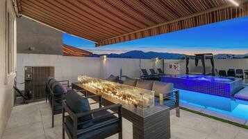 An outdoor patio with a fire feature, seating, a pool with blue lighting, and a view of mountains.
