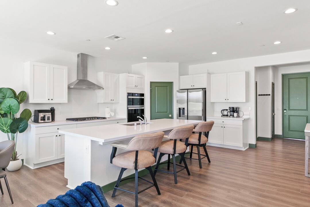 A modern kitchen with white cabinets, a central island with bar stools, a green door, and a stainless steel refrigerator.