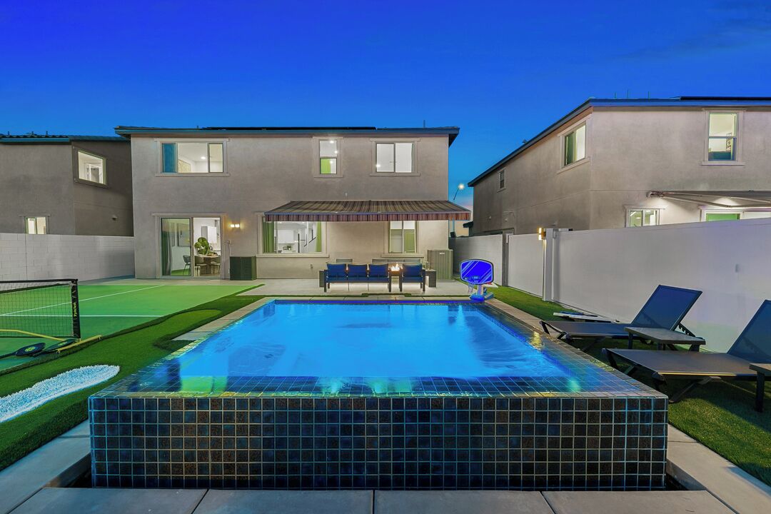 A swimming pool with a tiled deck, a covered patio area, and a view of a two-story house in the background.