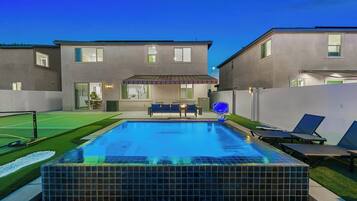 A swimming pool with a tiled deck, a covered patio area, and a view of a two-story house in the background.