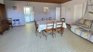Uma área de refeições com uma mesa posta para quatro pessoas, cadeiras de madeira, uma cozinha ao fundo e um sofá estampado.