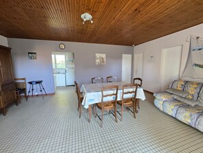 A dining area with a table set for four, wooden chairs, a kitchen in the background, and a patterned sofa. - Charming house near beach - 4 guests (Saint-Hilaire-de-Riez)
