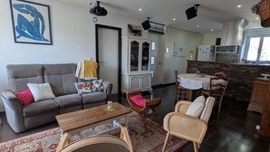 Een woonkamer met een bank, stoelen, een salontafel, een vloerkleed en een keuken met een eettafel.