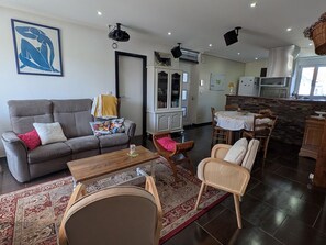 Ein Wohnzimmer mit einer Couch, Sesseln, einem Couchtisch, einem Teppich und einer Küchenzeile mit Esstisch.