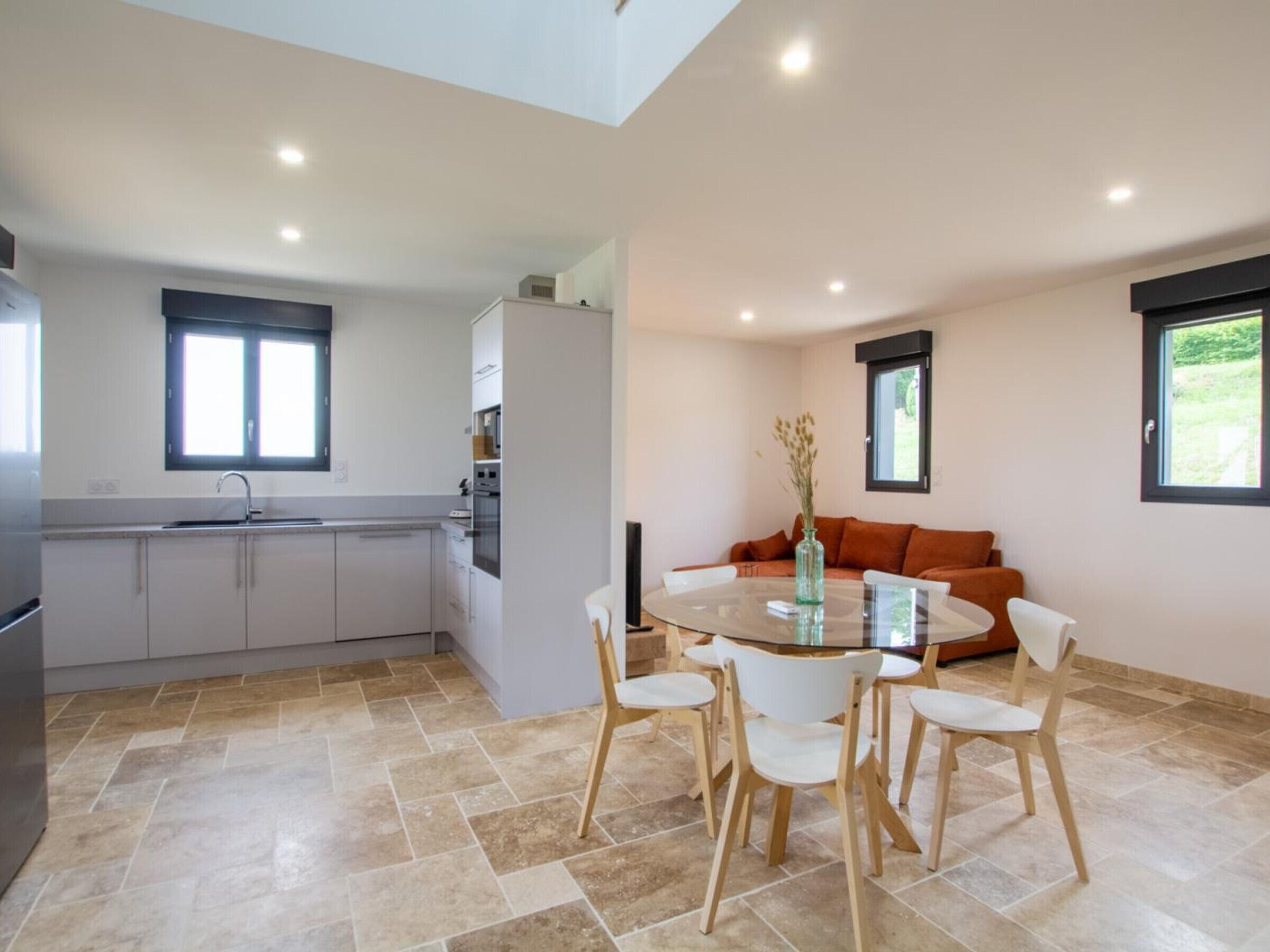 A modern kitchen with a dining area, featuring a round glass table, white chairs, and a sofa with orange cushions.