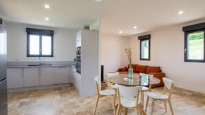 A modern kitchen with a dining area, featuring a round glass table, white chairs, and a sofa with orange cushions. - Single-storey house, pets allowed - 2 guests (Preyssac-d'Excideuil)