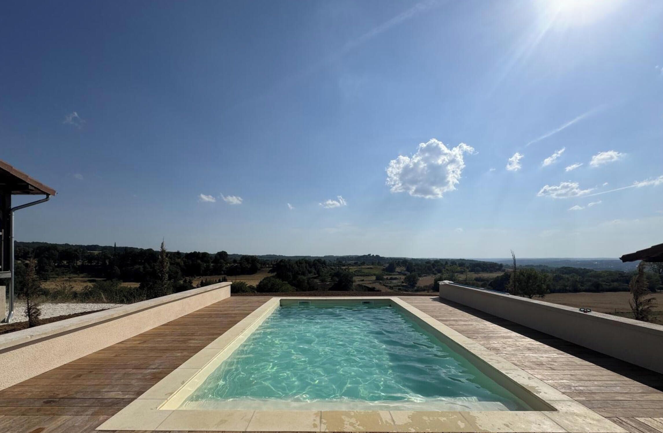 A rectangular swimming pool with a wooden deck surrounding it, set against a backdrop of a clear sky and distant landscape.