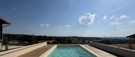 A rectangular swimming pool with a wooden deck surrounding it, set against a backdrop of a clear sky and distant landscape.