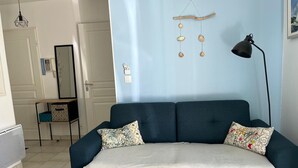 Ein modernes Wohnzimmer mit einem dunkelblauen Sofa, zwei gemusterten Kissen, einer hängenden Holzdeko, einer Stehlampe und einem kleinen Beistelltisch mit Spiegel und Dekorationsobjekten.