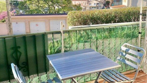 En balkon med et bord og to stole, gelænder i grøn metal og udsigt til bygninger og træer.