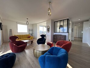 A modern living room with a sofa, armchairs, a coffee table, and a round ottoman. There is a view of the city through large windows. - Apartment sea view - 4 guests (La Rochelle)