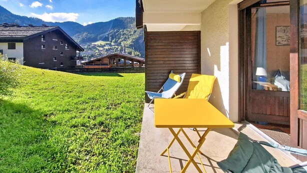 Un balcone con un tavolo giallo e sedie blu, affacciato su una zona verde e un paesaggio montano.