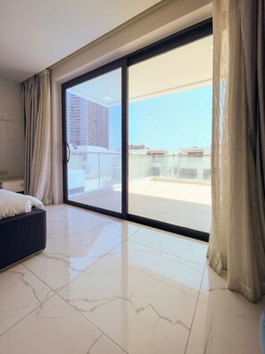 Una habitación de hotel moderna con un amplio ventanal corredero a un balcón, una cama y un piso de baldosas impecables.