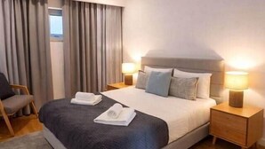 Ein modernes Hotelzimmer mit einem großen Bett, zwei Nachttischen inklusive Lampen, einem Sessel und Blick ins Freie durch das Fenster.