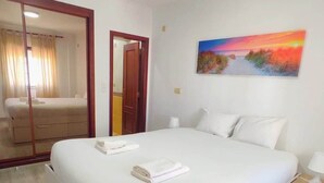 Une chambre d’hôtel avec un grand lit, une table de chevet, une commode, un tableau au mur et une porte donnant sur une autre pièce.